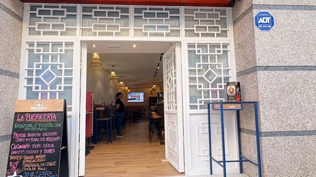 Fachada de La Puerkeria en Calle Azabache, 3, Carabanchel: puerta abierta con celosías blancas y la pizarra del menú del día